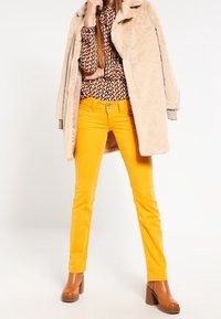 Abrigo de piel sintética beige, blusa estampada, pantalones amarillos de corte estrecho y botas de tacón marrones. El conjunto presenta una mezcla de texturas y colores vibrantes.