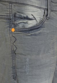 Šedé džíny vyrobené z denimu s oranžovými detaily, obsahující precizní design kapes a texturovanou tkaninu, vykazující jemné opotřebení a akcenty nití.