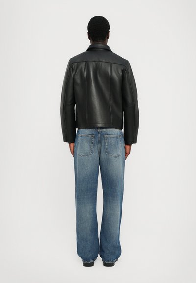 Chaqueta de cuero negra con hombros estructurados y cuello, combinada con pantalones vaqueros de pierna ancha en azul claro que cuentan con bolsillos traseros y un dobladillo sin terminar.