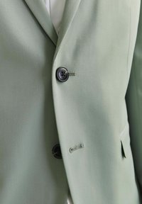 Chaqueta de traje verde claro hecha de una tela suave, con un solo botón negro y un sutil detalle de costura en la solapa.