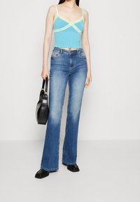 Ljusblå ribbad kort topp med gula spetsdetaljer, kombinerad med ljusblå utsvängda jeans och svarta platta skor, hållande en svart väska.