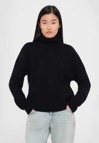 Navyfarbener Kabelstrickpullover mit hohem Rollkragen, gerippten Bündchen und Saum. Weiche Textur, entspannter Schnitt und auffällige Strickmusterdetails.