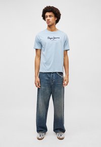 Ανοιχτό μπλε κοντομάνικο μπλουζάκι με μαύρο λογότυπο "Pepe Jeans London"; σε συνδυασμό με φαρδιά, ξεθωριασμένα μπλε τζιν και ασημένια αθλητικά παπούτσια.