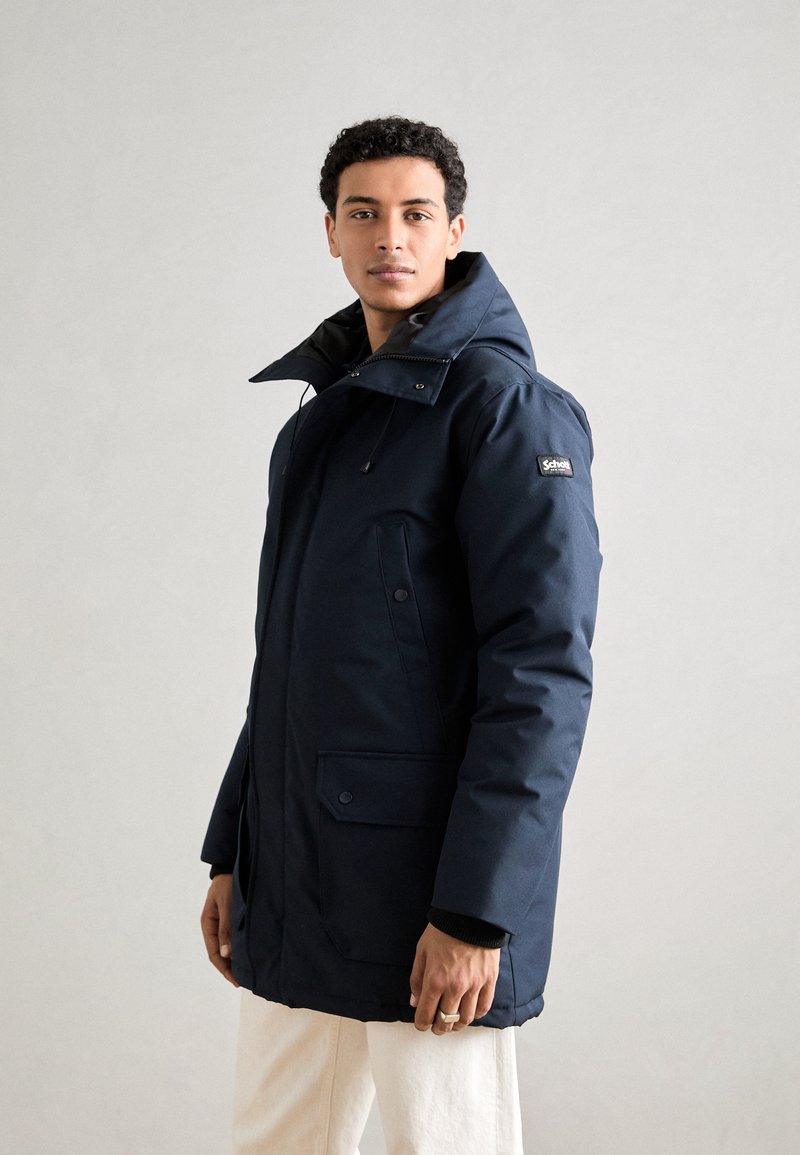 Navy blue waterproof parka with a high collar, front zip, and button pockets. Features drawstring hood and sleek, smooth texture.