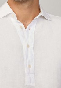 Camicia in lino bianco con abbottonatura a pressione con bottoni chiari, colletto appuntito e leggera trama visibile nel tessuto.