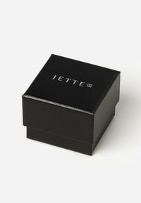 Svart smyckeslåda med texturerad yta och en silverfärgad "JETTE"-logotyp på toppen. Kompakt fyrkantig form med ett gångjärnslock.