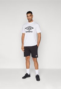 Camiseta blanca de manga corta con el logo negro de "Umbro", combinada con pantalones cortos negros que tienen el logo, zapatillas deportivas negras y calcetines de tipo crew blancos.
