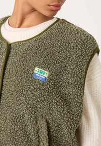 Žalias, pūkuotas liemenėlis, pagamintas iš tekstūruotos medžiagos su apvalia iškirpte, priekyje su mėlyna ir geltona etikete. Užsidedamas ant balto megztinio.