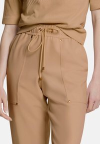 Beige joggerbroek met een elastische tailleband, een trekkoordsluiting, gouden accenten en twee voorzakken met drukknoopsluiting. Gladde stoftextuur.