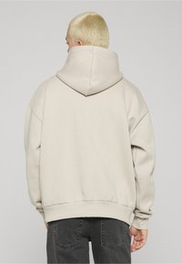 Bēšs pārmēru liels džemperis ar lielu kapuci, ribotiem aproču un apakšmalas maliņām; gluda tekstūra, bez rakstiem, valkāts kopā ar tumši pelēkām biksēm.