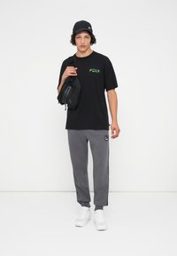 Melnā T-kreklā ar zaļu logo, pelēkās sporta biksēs ar sānu svītrām, melnā plecu somā un baltos sneakeros. Modelis stāv uz baltas fona.