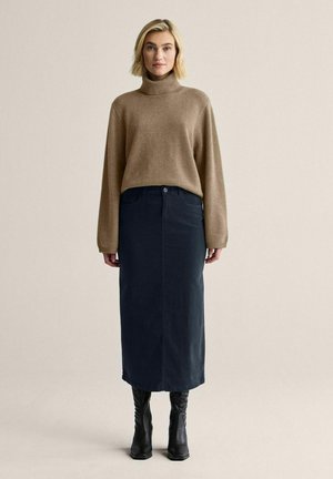 Lyshåret kvinde stående, iført en brun rullekravesweater, sort midi-nederdel og sorte knælange støvler mod en ensfarvet beige baggrund.