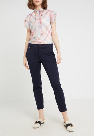 Femme portant un chemisier à manches courtes à carreaux pastel avec un nœud au col, un pantalon slim court bleu marine, et des chaussures blanches en slip-on avec des brides noires.