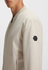 Hellbeige Bomberjacke mit hexagonalem Muster, geripptem Kragen und schwarzem Sechseck-Logo am Ärmel. Glatte Textur mit strukturiertem Design.