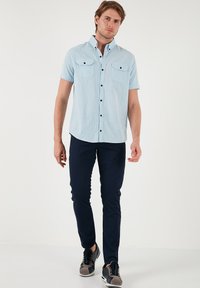 Camisa corta de manga azul clara con dos bolsillos en el pecho y botones negros, combinada con pantalones marinos y zapatillas gris. Textura de tela suave.