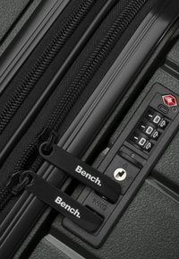 Detalles de la cremallera de una maleta negra con tiradores de doble marca y un candado de combinación, sobre una superficie texturizada en gris.