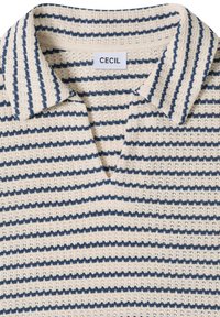 Polo en maille beige avec des rayures horizontales bleu marine et une étiquette blanche portant l'inscription "CECIL" à l'arrière du col intérieur.