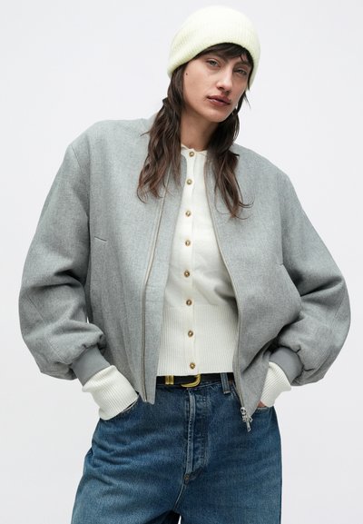 Veste bomber grise avec poignets élastiques, portée sur un cardigan crème boutonné et un jean taille haute bleu. Le mannequin porte un bonnet jaune clair.