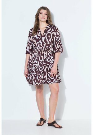 Kobieta ubrana w luźną sukienkę do kolan w brązowo-biały wzór, czarne sandały na koturnie, stojąca na białym tle.