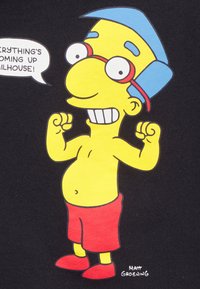 Piirretty hahmo, jolla on keltainen iho, punaiset shortsit ja kengät, sininen hattu ja silmälasit; hymyilee nyrkit nostettuina. Musta tausta.