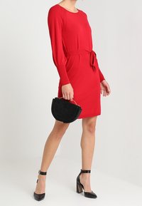 Robe tricotée rouge à manches longues, taille ceinturée et poches latérales. Associée à des escarpins noirs et un sac à main rond noir. Texture lisse.