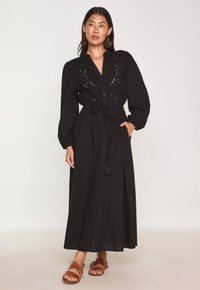 Robe maxi noire avec des broderies, fermeture à boutons sur le devant, manches ballons, taille élastique avec une ceinture nouée, et poches latérales.
