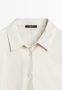 Chemise col crème avec deux boutons visibles et une étiquette noire "Massimo Dutti" à l'intérieur de l'encolure.