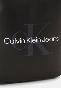 Borsa in pelle nera con un prominente logo bianco "Calvin Klein Jeans" e un grande emblema "CK" in un delicato font grigio. Texture liscia.