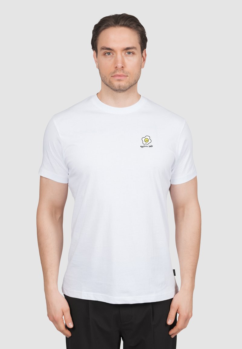 Homme aux cheveux courts bruns portant un t-shirt blanc avec un petit motif d'œuf au plat souriant et le texte "eggstra smile" sur le côté gauche de la poitrine.