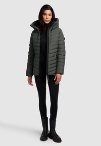 Grüne Puffjacke mit hohem Kragen und Reißverschluss, ausgestattet mit einem steppierten Design. Getragen über einem schwarzen Outfit mit eng anliegenden Hosen und schwarzen Stiefeln.