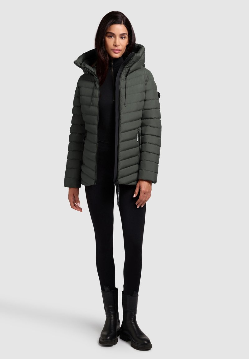 Grüne Puffjacke mit hohem Kragen und Reißverschluss, ausgestattet mit einem steppierten Design. Getragen über einem schwarzen Outfit mit eng anliegenden Hosen und schwarzen Stiefeln.