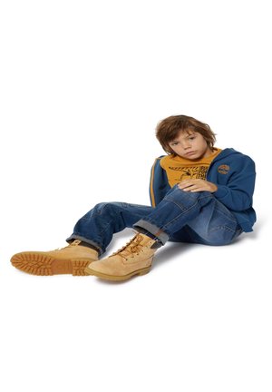 Tenue d'enfant : veste zippée bleue, t-shirt jaune avec un design imprimé, jeans en denim et bottes à lacets beiges avec semelles en caoutchouc et motif texturé.