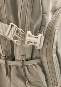 Boucle de libération latérale en plastique fixant deux sangles beiges sur un tissu beige avec des fermetures éclair et des cordons visibles.