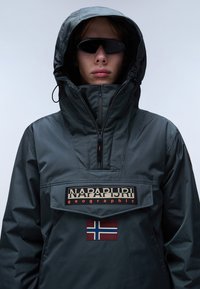 Tummanharmaa pullover-takki, jossa on korkea kaulus, etuvetoketju, logopatch ja norjalaisen lipun yksityiskohta vartalolla. Varustettu suurella, säädettävällä hupulla.