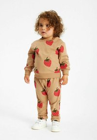 Completo in cotone beige composto da felpa e pantaloni con stampe di fragole rosse. Materiale morbido, polsini e vita a costine con una vestibilità elastica. Sneakers bianche.