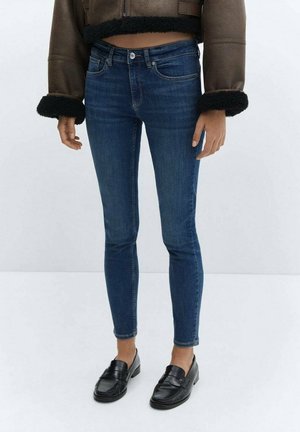 Person trägt dunkelblaue Skinny-Jeans, schwarze Loafers und eine braune kurze Jacke mit schwarzem Pelzbesatz an Ärmeln und Saum.