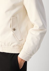Light cream bomber jacket with ribbed hem and cuffs, featuring a side pocket with a button detail, smooth texture, and sporty design.