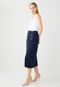 Gonna midi blu navy realizzata in tessuto elasticizzato, con dettagli in bottoni dorati e uno spacco laterale, abbinata a una canotta bianca e tacchi beige.
