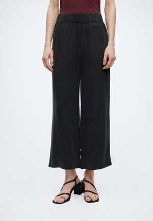 Žena oblečená v voľných čiernych širokých krátkych nohaviciach s elastickým pásom a čiernych sandáloch na blokovom opätku so strapcami, stojaca na bielom pozadí.