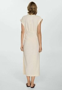 Vrouw die met haar rug naar de camera staat, in een beige mouwloos midi-jurkje en zwarte instap sandalen, tegen een effen lichte achtergrond.