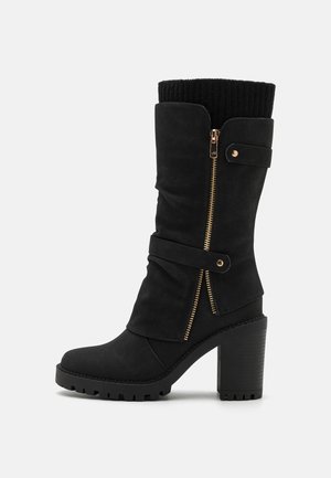 Botte noire mi-mollet à talon avec semelle épaisse, fermeture éclair latérale dorée, sangles décoratives et bord-côte tricoté sur fond clair.