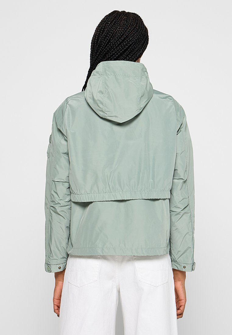 Hellgrünes, kurzes Jacket aus glattem, leichtem Material mit Kapuze. Verfügt über einen elastischen Bund und Druckknopfakzente an den Ärmeln.