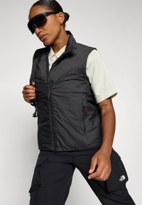 Gilet isolato nero con zip frontale, tasche laterali e colletto alto, abbinato a una camicia leggera e pantaloni cargo. Grandi occhiali da sole scuri.