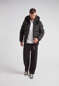 Giubbotto puffo nero con cappuccio, dotato di tasche con zip e una toppa con logo bianco. Abbinato a pantaloni della tuta neri e sneaker.