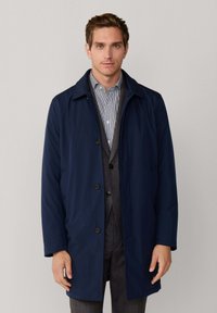 Jonge man in een marineblauwe jas over een grijs pak en gestreept shirt, staand tegen een effen lichtgrijze achtergrond.