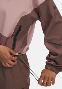 Hände, die den Kordelzug einer rosa und braun farblich abgesetzten Jacke mit Reißverschlusstasche und elastischem Bund am Ärmel anpassen.