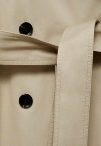 Beige trenchcoatstof met een gestructureerde afwerking, met een zwarte knop en een bijpassende riem die om de taille is geknoopt.
