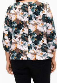 Blusa de estampado floral con un corte relajado, mangas de tres cuartos y un discreto detalle de abertura en la parte posterior. Los colores incluyen verde azulado, marrón y naranja.