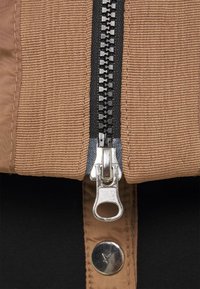 Primo piano di una giacca di stoffa marrone caratterizzata da una zip nera e hardware argentato, con costine testurizzate lungo il tessuto. Fodera nera visibile.