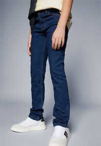 Jeans de denim albastru închis cu o croială slim, av�ând un design clasic cu cinci buzunare și cusături vizibile, asortați cu adidași albi.